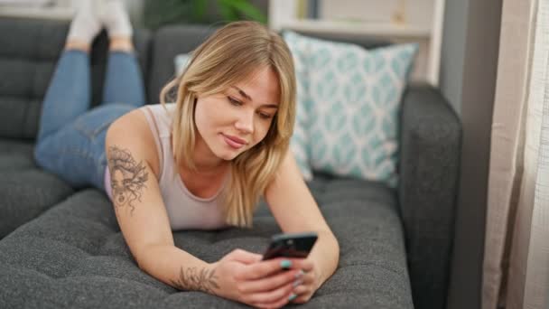 年轻的金发女人用智能手机躺在沙发上 — 图库视频影像