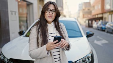Genç, güzel, İspanyol bir kadın akıllı telefon kullanıyor. Arabasının yanında duruyor.