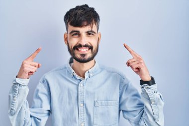 Mavi arka planda duran sakallı genç İspanyol adam iki eliyle başını işaret ederek gülümsüyor, harika bir fikir ya da düşünce, iyi bir hafıza 