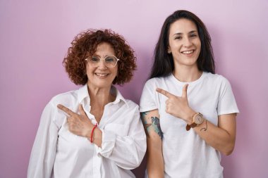 İspanyol anne ve kızı pembe arka plan üzerine günlük beyaz tişört giyiyorlar. Neşeli bir şekilde gülümsüyorlar. Yüzlerinde mutlu ve doğal bir ifade var. 