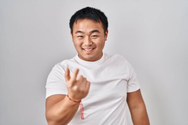 Beyaz arka planda duran genç Çinli adam seni buraya davet ediyor. El kol hareketi yap. Mutlu bir şekilde karşıla ve gülümse. 