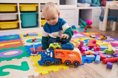 Beyaz tenli sevimli bir çocuk anaokulunda yerde oturmuş oyuncaklarla oynuyor.