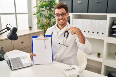 Doktor steteskobu takan genç İspanyol adam elinde dosya tutarak gülümseyen mutlu ve gururlu birini işaret ediyor. 