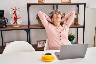 Orta yaşlı kadın, elleri başının üstünde evde masada otururken rahatlıyor.