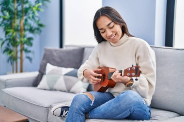 Evdeki kanepede oturan genç İspanyol kadın ukulele oynuyor.