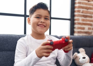 Adorable hispanic boy playing video game sitting on sofa at home