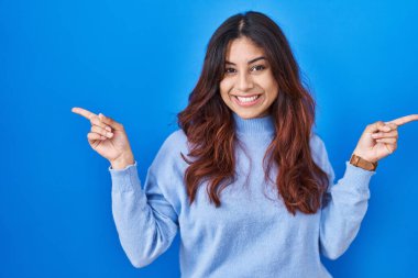 Mavi arka planda duran Latin kökenli genç bir kadın parmaklarıyla farklı yönleri işaret ederek gülümsüyor. reklam için kopyalama alanı 