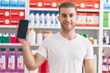 Eczanede çalışan beyaz bir genç akıllı telefon ekranı gösteriyor. Pozitif ve mutlu görünüyor. Kendine güvenen bir gülümsemeyle dişlerini gösteriyor. 