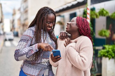 Afro-Amerikalı kadın arkadaşlar, sokakta akıllı telefon kullanarak gülümsüyorlar.