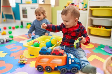 Anaokulunda yerde oturan top ve araba oyuncaklarıyla oynayan sevimli çocuklar.