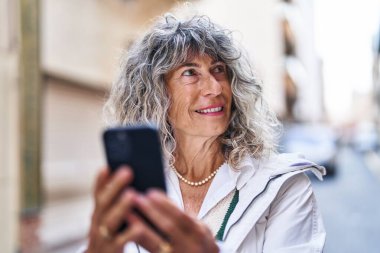 Sokakta akıllı telefon kullanan orta yaşlı bir kadın gülümsüyor.