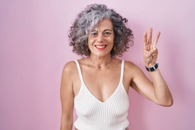 Middle age woman with grey hair standing over pink background showing and pointing up with fingers number three while smiling confident and happy. 