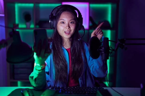 若いアジアの女性がカメラを見て側に手指で指して 顔に大きな笑顔でスマートフォンでビデオゲームをプレイ — ストック写真