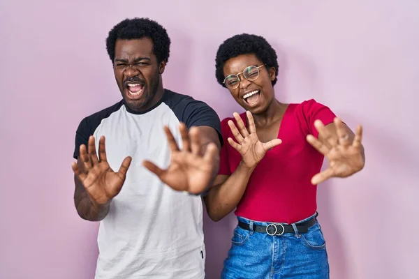 年轻的非洲裔美国夫妇站在粉红的背景上 害怕而且害怕 恐惧的表情 用手停止手势 震惊地大叫 恐慌概念 — 图库照片