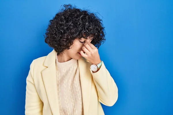 年轻的黑发女人 卷曲的头发站在蓝色的背景上 疲倦地揉揉鼻子和眼睛 感到疲倦和头痛 压力和挫败感概念 — 图库照片