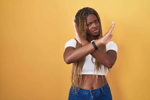 African american woman with braided hair standing over yellow background rejection expression crossing arms doing negative sign, angry face