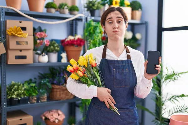 Brunette woman working at florist shop holding smartphone puffing cheeks with funny face. mouth inflated with air, catching air.