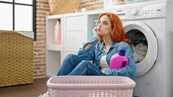 在洗衣房 年轻的红头发女人靠着洗衣机 手里拿着洗涤剂瓶子 疲惫不堪 — 图库照片