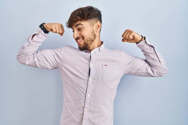 Arab man with beard standing over blue background showing arms muscles smiling proud. fitness concept. 