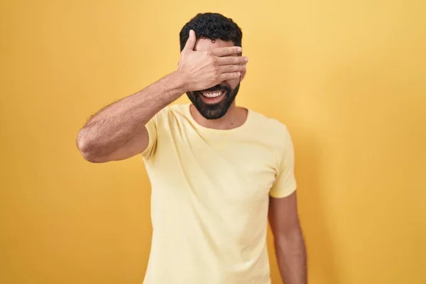 黄色い背景の上に髭を生やしたヒスパニック系の男性が笑顔で 驚きのために目を覆う顔で手で笑っています 視覚障害の概念 — ストック写真