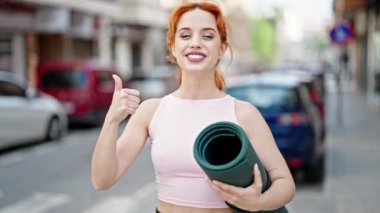 Elinde yoga minderiyle gülümseyen genç kızıl saçlı kadın sokakta el hareketi yapıyor.