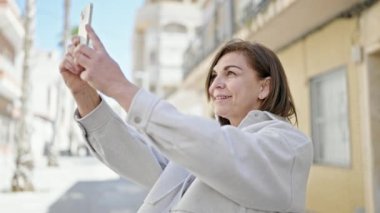 Orta yaşlı, kendine güvenen İspanyol bir kadın sokakta akıllı telefonuyla fotoğraf çekiyor.