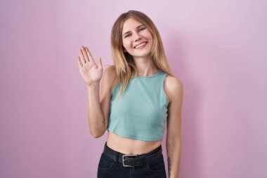 Blonde caucasian woman standing over pink background waiving saying hello happy and smiling, friendly welcome gesture 