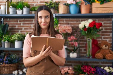 Young beautiful hispanic woman florist smiling confident reading notebook at flower shop