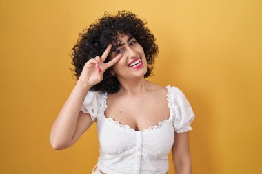 Sarı arka planda duran kıvırcık saçlı genç esmer kadın barış sembolü yapıyor. Yüzünde parmakları, gülümseyerek zaferini gösteriyor. 