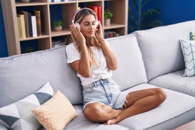 Young blonde woman listening to music sitting on sofa at home
