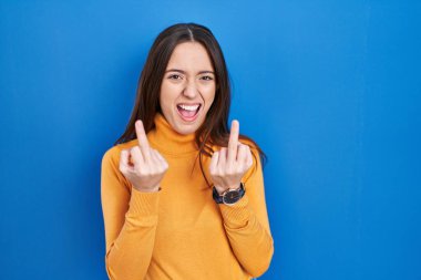 Young brunette woman standing over blue background showing middle finger doing fuck you bad expression, provocation and rude attitude. screaming excited 