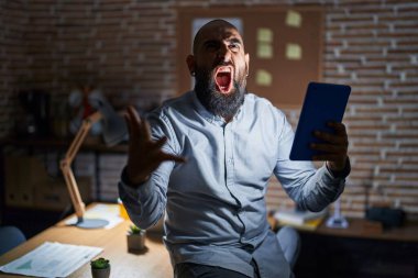 Geceleri ofiste çalışan sakallı ve dövmeli genç İspanyol bir adam sinirli ve öfkeli bağırıyor ve öfkeyle bağırıyor. öfke ve agresif konsept. 