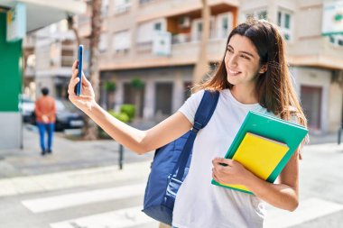 Young beautiful hispanic woman student holding books make selfie by smartphone at street