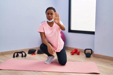 Spor kıyafetleri ve güvenlik maskesi takan genç Afro-Amerikalı spor salonunda dur işareti yapıyor ciddi ve kendinden emin bir ifadeyle savunma jesti yapıyor. 