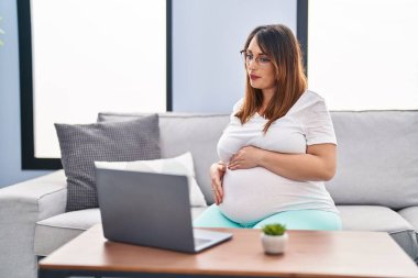 Genç hamile bir kadın evdeki koltukta video görüşmesi yapıyor.