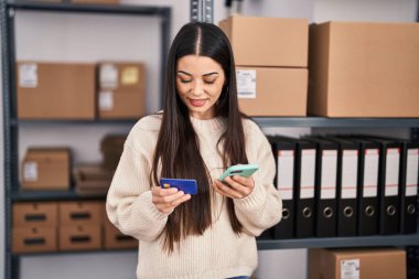 Young beautiful hispanic woman ecommerce business worker using smartphone and credit card at office