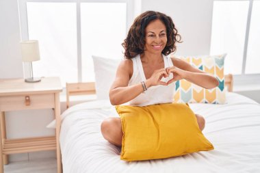 Middle age woman doing heart gesture sitting on bed at bedroom