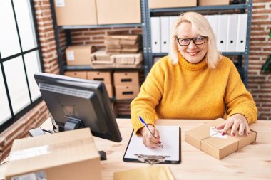 Middle age blonde woman ecommerce business worker writing on document working at office