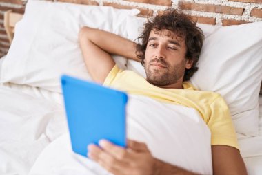 Young hispanic man watching video on touchpad lying on bed at bedroom