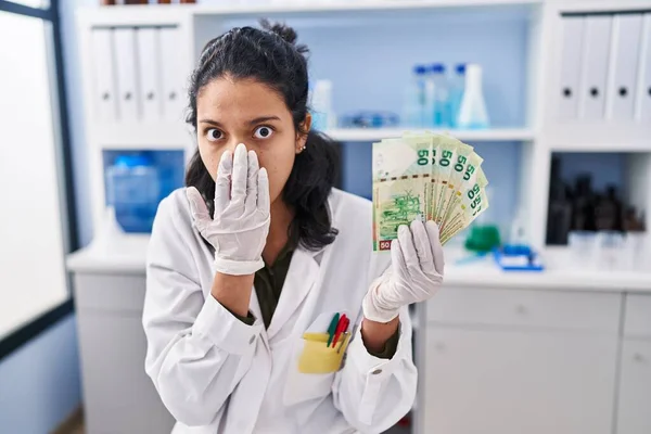 暗い髪を持つヒスパニック系の女性は 手で口をカバーお金を保持科学研究所で働いています ショックを受け 間違いを恐れて 驚きの表情 — ストック写真