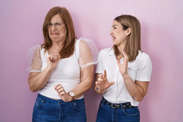 Hispanic mother and daughter wearing casual white t shirt over pink background disgusted expression, displeased and fearful doing disgust face because aversion reaction.