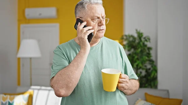 中年白髪の男飲むコーヒー話して上のスマートフォンで自宅 — ストック写真