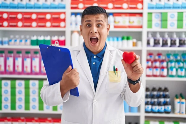 Joven Hombre Hispano Trabajando Farmacia Sosteniendo Corazón Celebrando Loco Sorprendido —  Fotos de Stock