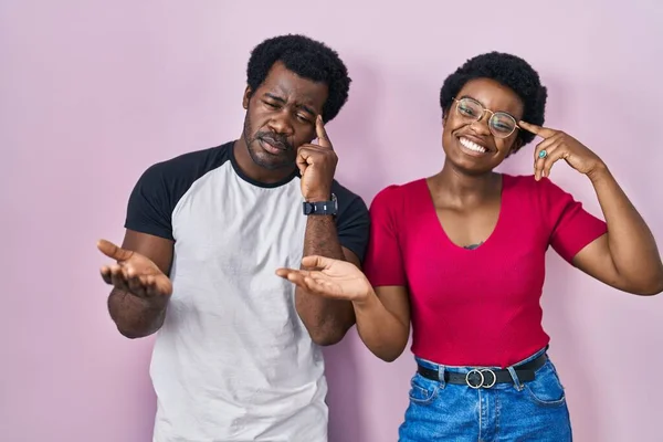 年轻的非洲裔美国夫妇站在粉红色的背景上 对张开的手掌展示复制的空间和指尖指向额头感到困惑和烦恼 好好想想吧 — 图库照片