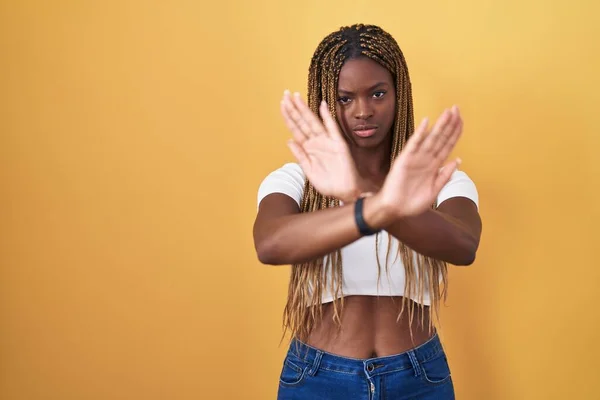 African american woman with braided hair standing over yellow background rejection expression crossing arms and palms doing negative sign, angry face