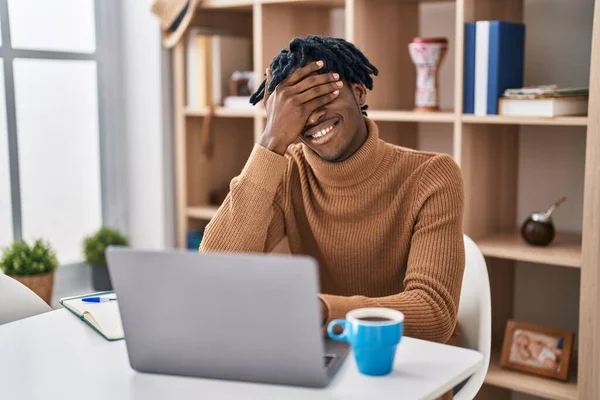 Young african man with dreadlocks working using computer laptop smiling and laughing with hand on face covering eyes for surprise. blind concept.