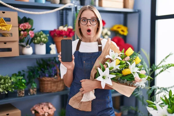 スマートフォンの画面を示す花屋で働く若い白人女性は恐れていると驚きと驚きの表情でショックを受けました 恐怖と興奮した顔 — ストック写真
