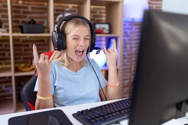 年轻的高加索女人带着耳机玩电子游戏 带着疯狂的表情大喊着举起手做摇滚符号 音乐明星 重音乐概念 — 图库照片
