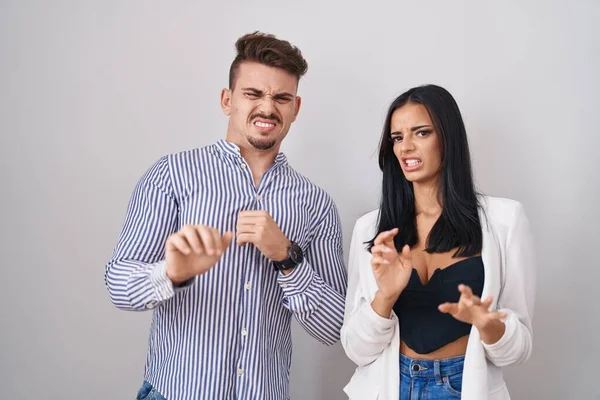 Young hispanic couple standing over white background disgusted expression, displeased and fearful doing disgust face because aversion reaction.