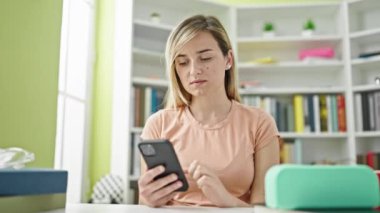 Kütüphanede akıllı telefon kullanan genç sarışın kız öğrenci.